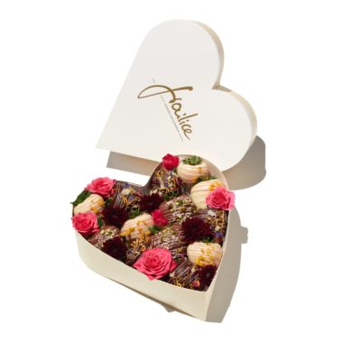 Eine Box in Herzform, gefüllt mit bunt verzierten Erdbeeren sowie rosa, pinken und roten Rosen.