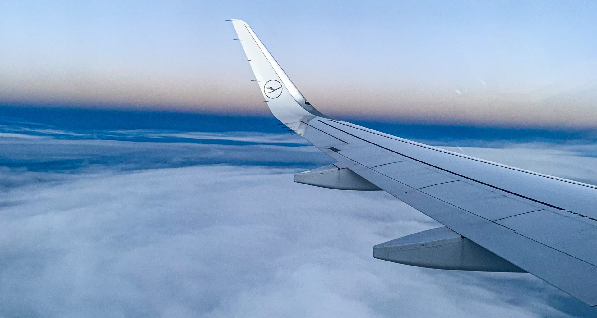Erster Flug „nach“ Corona: Der Flügel eines Lufthansa-Flugzeugs im Vordergrund, Wolken und ein Sonnenuntergang im Hintergrund.