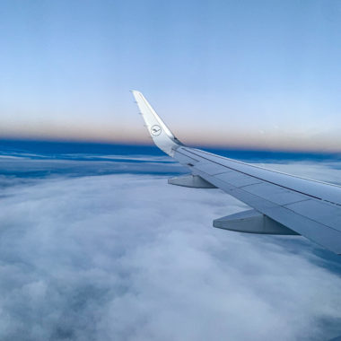 Erster Flug „nach“ Corona: Der Flügel eines Lufthansa-Flugzeugs im Vordergrund, Wolken und ein Sonnenuntergang im Hintergrund.