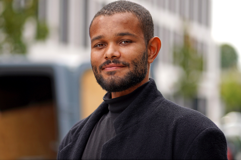 Portrait einer männlichen Person of Color mit kurzen Haaren in einem schwarzen Pullover und schwarzem Wollmantel in einer urbanen Umgebung